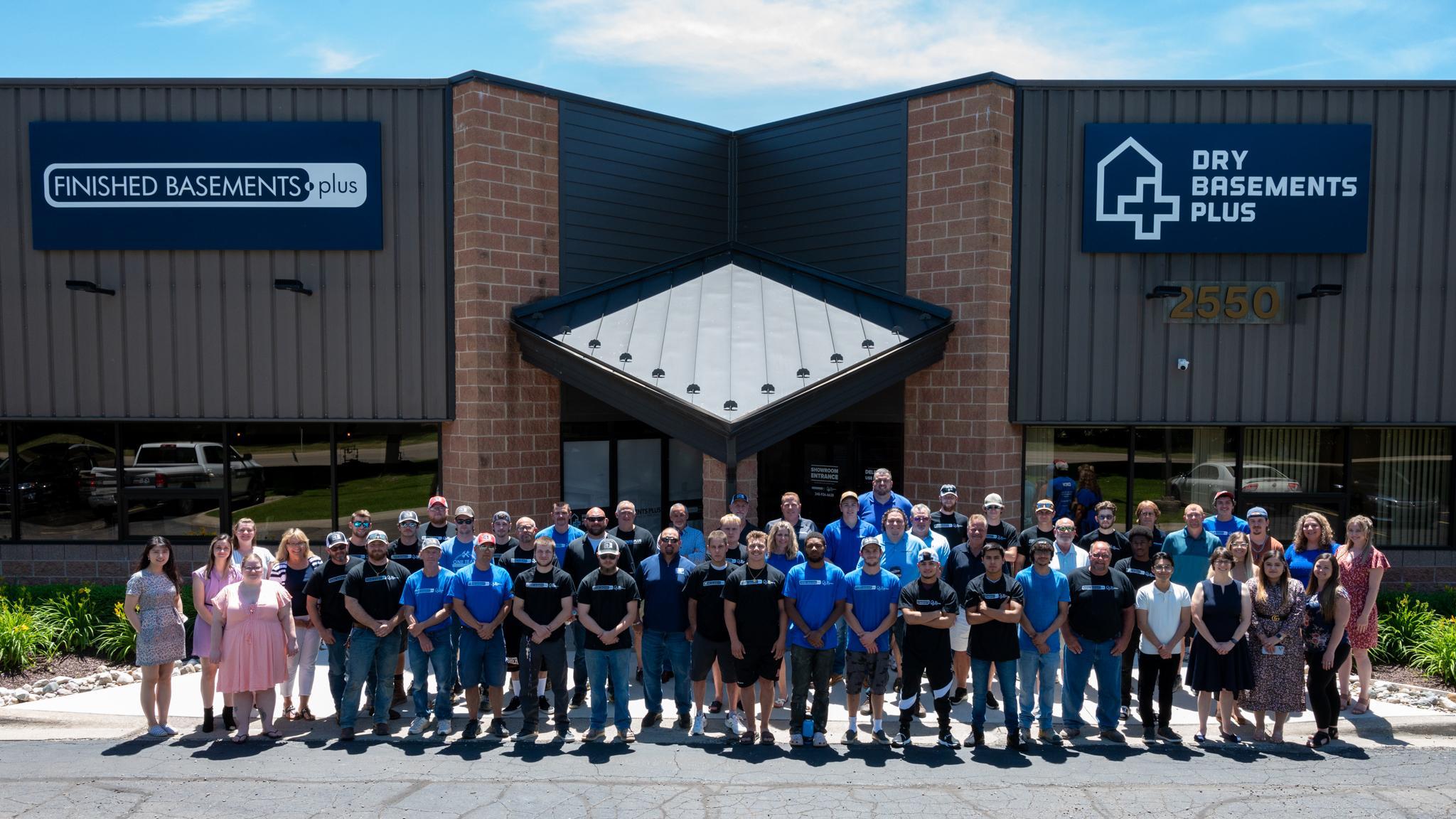 Photo of the Finished Basements and Dry Basements Plus office with brick and dark grey paneling. Employees standing in front of the building. Photo taken from a raised position.