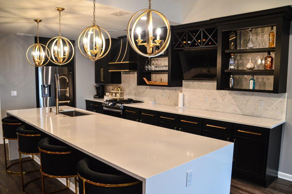 Kitchen area with long white counter island, black cabinets over the back counter, and gold hanging lights.