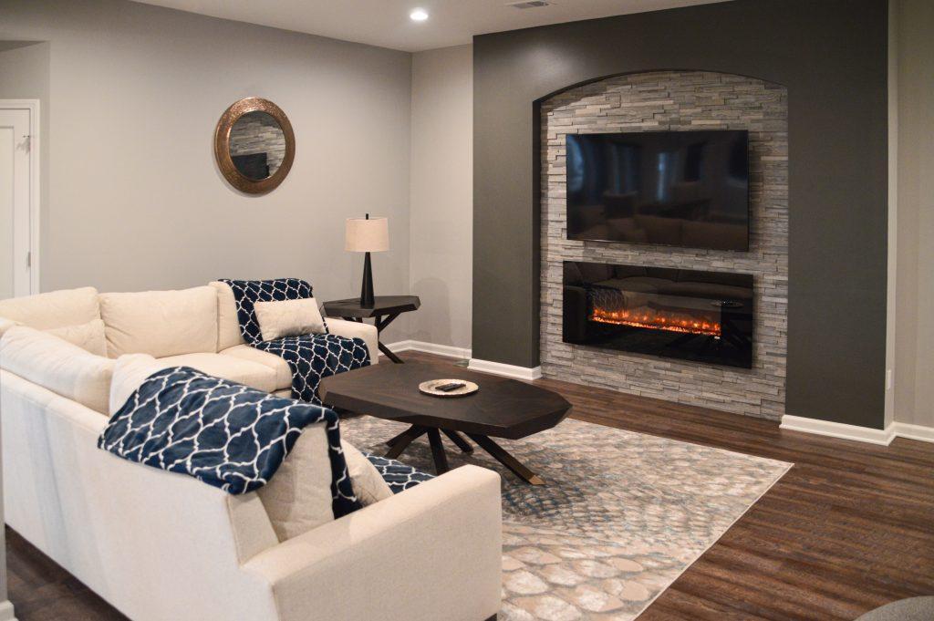 Finished basement Livingroom area with sectional couch, fireplace under a TV over a stone backsplash.