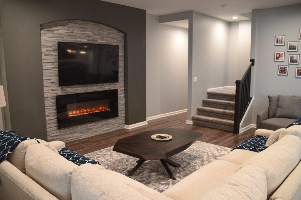 Finished basement Livingroom area with sectional couch, fireplace under a TV over a stone backsplash.