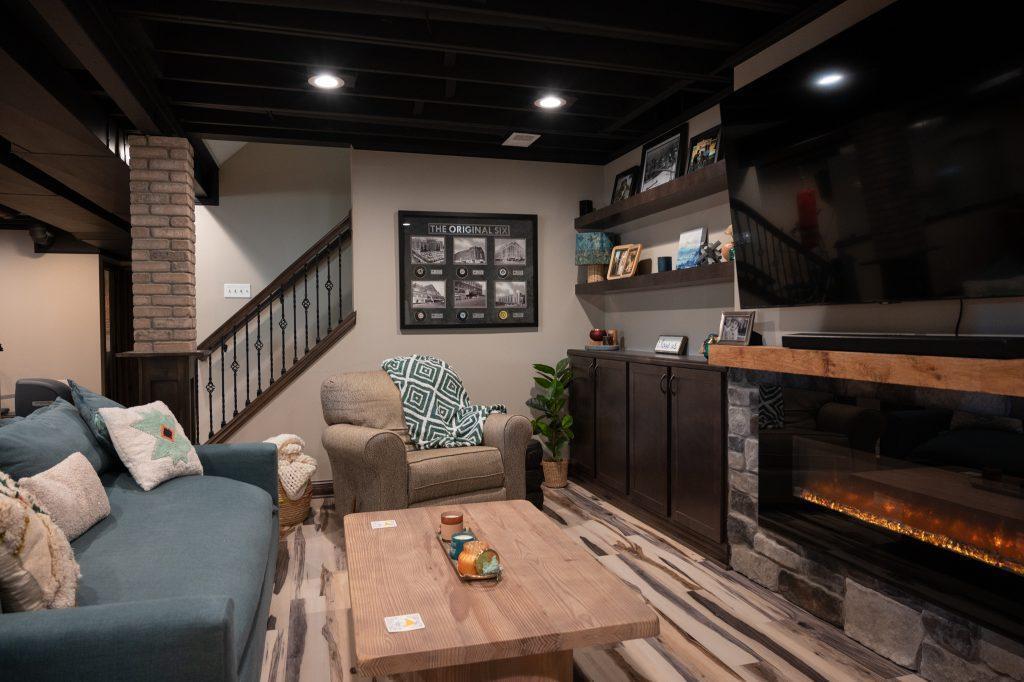 Finished basement living room area looking towards the stairs in the back along with a view of the tv with fireplace underneath and built-in shelving unit displaying family photos.