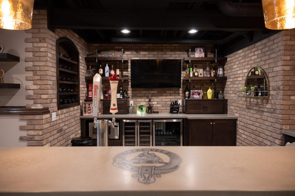 Finished basement home bar view from the middle of the custom cement bar top looking at the back of the bar with shelving for spirits and tv in the middle.