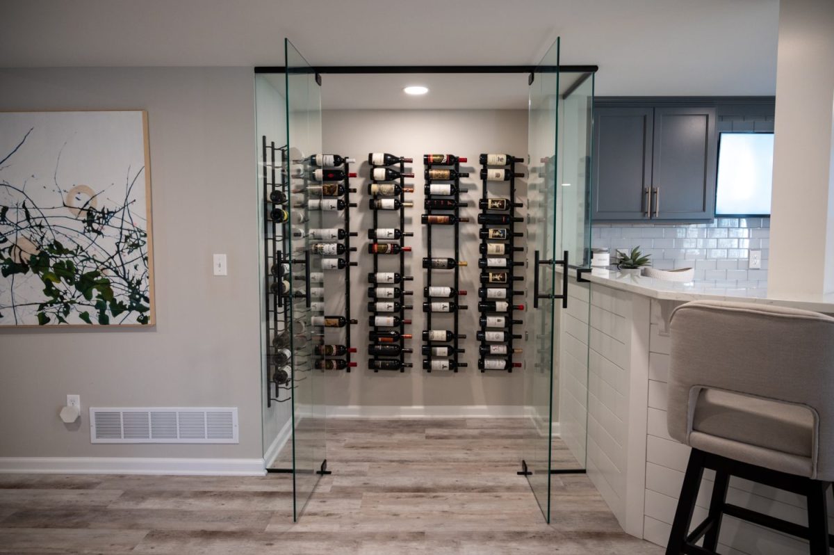 Photo of glass-encased wine storage room next to a white tile kitchenette and bar. Basement remodeled by Basements Plus. Northville, Michigan