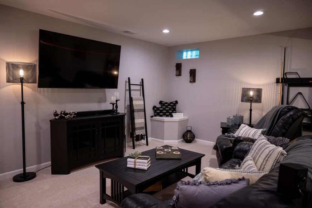 Photo of Finished Basements remodeled basement. Light colored walls with black leather and dark wood furnishings. Modern lighting with big screen TV mounted on the wall.