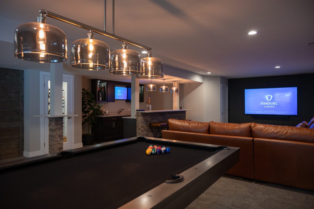 Photo of Basements Plus remodeled basement with brown leather couch, large-screen TV mounted on dark accent wall, kitchenette with brick bar, and industrial lighting above a dark pool table. Grand Rapids, Michigan.