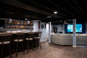 Finished basement "man-cave" with wood floor. A large, angled white couch sits in front of a large screen TV mounted on a dark blue accent recess. A bar with modern hanging lights and wooden barstools to the left. Various bottles of alcohol sit on mounted dark wood shelves. Southfield, Michigan. Remodeled by Basements Plus.