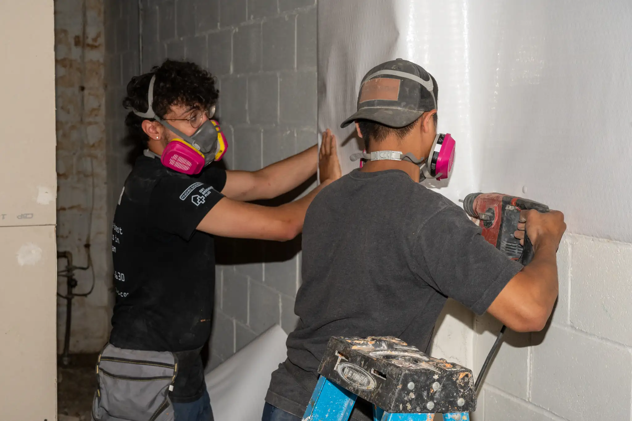 Two Basements Plus crew members wearing pink/yellow/gray respirators as their cover a basement wall to secure against moisture intrusion.